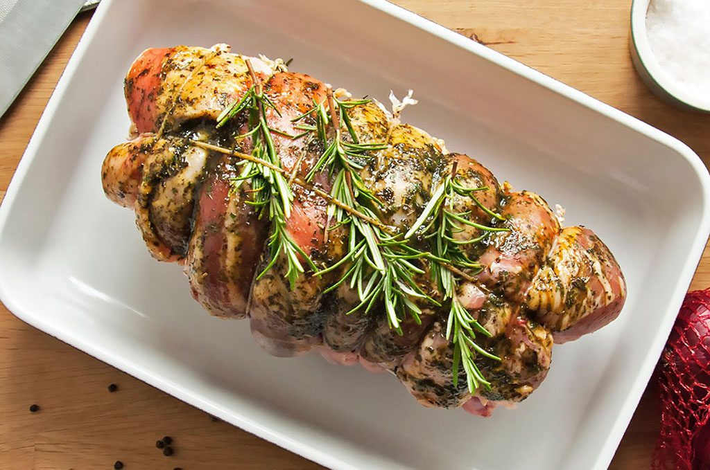 A roast lamb roll, sitting in a white baking dish with a sprig of rosemary